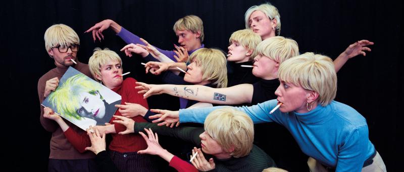 Eine Gruppe junger Menschen mit weißblonden Perücken recken sich nach einem Bild von Andy Warhol
