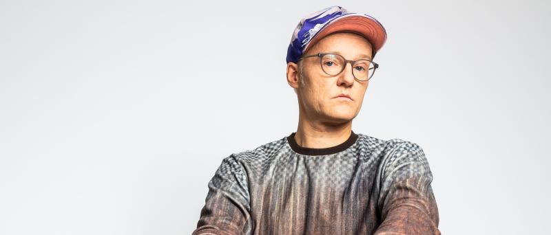 Peter Licht sitzt vor einem neutralen Hintergrund. Er trägt ein graues Oberteil, eine Brille und eine bunte Cap.
