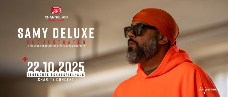 Der Künstler Samy Deluxe in orangem Pullover, roter Mütze und dunkler Sonnenbrille vor einem hellen, leicht unscharfen Hintergrund.
