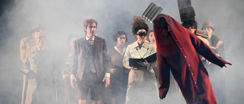 Bühnensituation: Das Ensemble steht als Gruppe zusammen in zwei Reihen. Sie tragen lange braune und beige Trenchcoats und halten Bücher in der Hand. Eine Person balanciert einen Stapel Bücher auf dem Kopf und der Schulter. Sie schauen auf eine Figur im Vordergrund. Sie trägt ein körperlanges rotes zugeknöpftes Hemd mit langen Ärmeln, das weit oberhalb des Kopfes endet, sodass kein Kopf zu sehen ist. Die Figur steht schräg, ebenfalls mit einem schiefen Stapel Bücher auf dem Kopf. Die Szenerie ist in dichten 