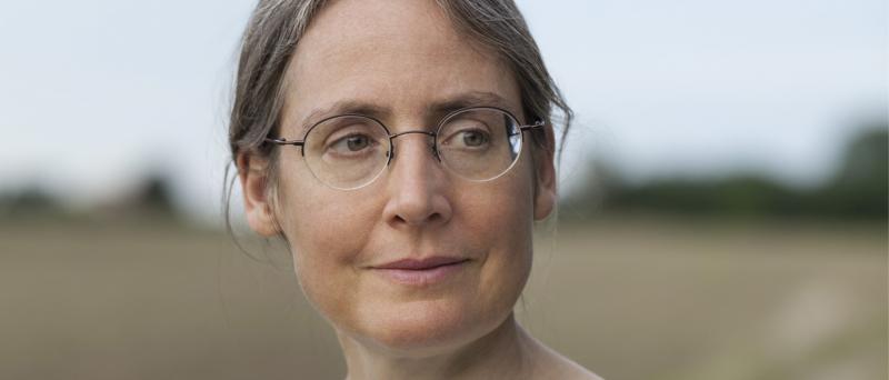 Porträt. Isabel Fargo Cole steht draußen in der Natur. Sie lächelt ganz leicht und schaut an der Kamera vorbei. Die trägt zusammengebundenes Haar und eine filigrane Brille.