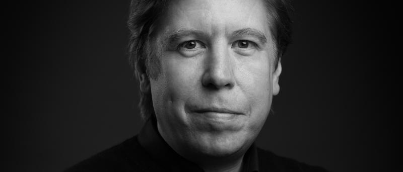 Schwarz-Weiß-Foto. Benjamin Sprick steht vor einem neutralen Hintergrund. Er trägt ein dunkles Hemd und schaut in die Kamera. Er schmunzelt leicht.