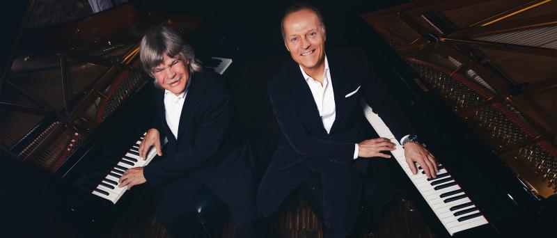 Die Pianisten Axel Zwingenberger und Joja Wendt sitzen jeweils an einem Flügel. Die Kamera ist in der Draufsicht leicht von oben platziert. Beide tragen ein weißes Hemd und einen dunklen Anzug. Sie lächeln in die Kamera.