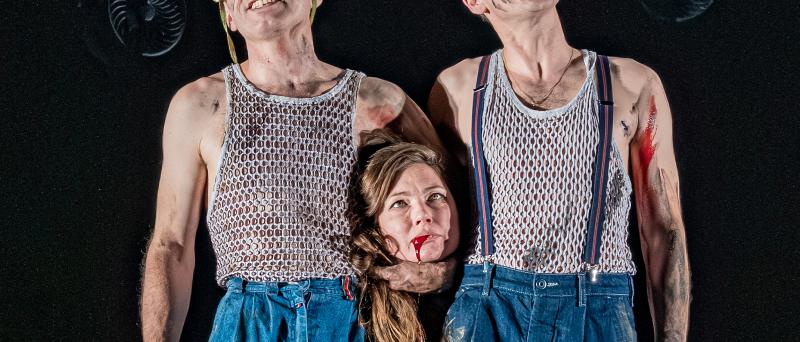 Kristof Van Boven und Nils Kahnwald stehen nebeneinander auf der Bühne. Sie tragen dreck- und blutverschmierte blaue Jeanshosen und graue Unterhemden mit vielen kleinen Löchern. Beide lächeln, unter dem Arm von Kristof Van Boven ist der Kopf von Sabine Molenaar zu sehen, ihr Haar fällt herunter, sie schaut nach oben, aus ihrem Mund läuft Blut. Über ihnen hängt eine große runde Stahlkonstruktion aus vielen unterschiedlich großen alten trichterförmigen Lautsprechern, Scheinwerfern und Ventilatoren. 