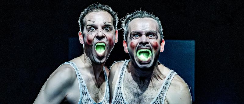 Nils Kahnwald und Kristof Van Boven nebeneinander in Nahaufnahme. Sie tragen weiße Unterhemden mit vielen kleinen Löchern und schauen mit verzerrtem Gesicht in die Kamera. Ihre Wangen sind grob mit roter Farbe bemalt, die Nase und Augenbrauen von Nils Kahnwald sind mit schwarzem Stift grob skizziert. Ihre Münder sind weit aufgerissen, es leuchtet grellgrün aus ihnen.