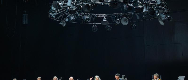 Alle sieben Zeitzeug*innen der Inszenierung sitzen nebeneinander auf der Bühne. Eine Person mittig hat eine Hand gehoben und Mittel- und Zeigefinger gespreizt. Alle schauen zu ihr. Rechts steht Julia Wieninger an einem Mikrofon, Kristof Van Boven sitzt zu den Füßen der sprechenden Person und schaut sie an. Über der Szene hängt eine große runde Stahlkonstruktion aus vielen unterschiedlich großen alten trichterförmigen Lautsprechern, Scheinwerfern und Ventilatoren.