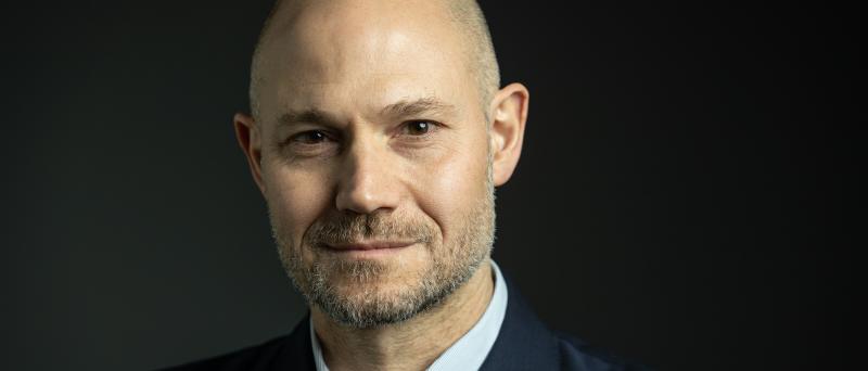 Omri Boehm steht vor einem dunklen, neutralen Hintergrund. Er trägt einen dunklen Anzug und lächelt in die Kamera.