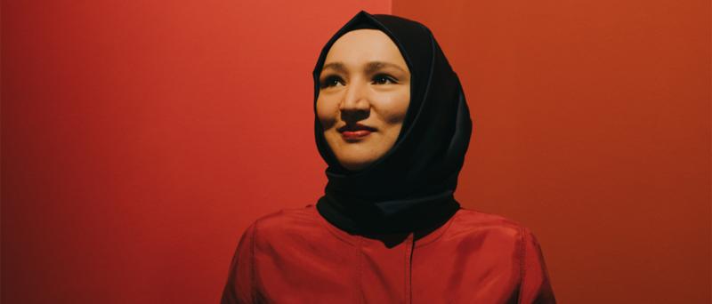 Kübra Gümüşay steht vor einem roten Hintergund. Die trägt ein rotes Oberteil und ein schwarzes Kopftuch. Sie lächelt und schaut leicht an der Kamera vorbei.