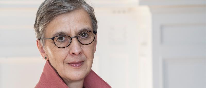 Katharina Pistor steht vor einem neutralen Hintergrund. Sie trägt kurze, graue Haare, eine dunkle Brille und ein rosafarbenes Oberteil. Sie schaut in die Kamera.