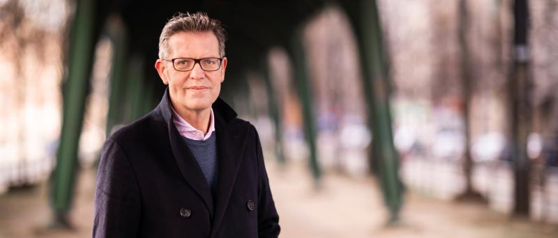 Steffan Mau steht draußen unter einer Brücke, die aussieht, wie eine Hochbahnbrücke. Er trägt eine dunkle Jacke, kurze graue Haare und eine Brille. Er schaut in die Kamera.