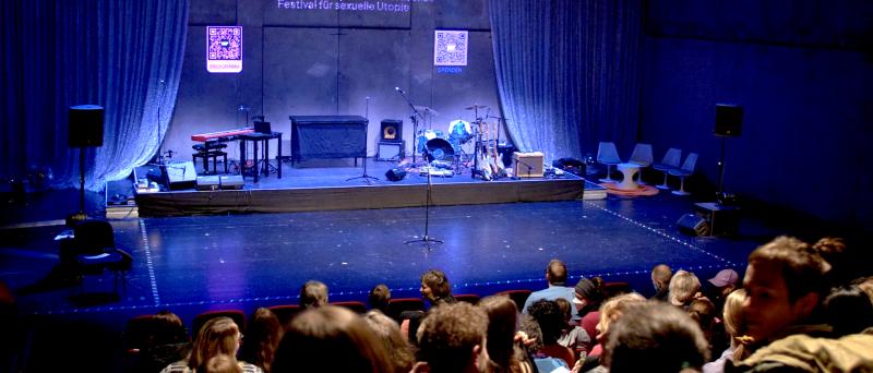 Eine Bühne ist mit Musikinstrumenten eingerichtet. Rechts und links von der Bühne hängt ein heller Vorhang. Über der Bühne ist der Schriftzug "SUPERZART* - intersektionales queerfeministisches Festival für sexuelle Utopie" zu lesen. Vor der Bühne sitzen viele Menschen auf roten Theatersitzen.