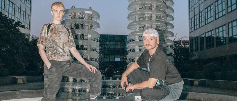 lynn t musiol steht an einem Wasserspiel-Brunnen und hat ein Bein auf dem Rand des Brunnens abgestellt. Neben ihr hockt Marcus Peter Tesch auf dem Rand des Brunnens. Vor den beiden steht eine steinerne Sitzbank.