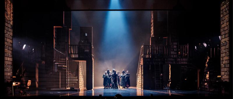 Bühnensituation. Die Bühne ist in der Totalen zu sehen. Rechts und links stehen riesige Bücherregale. In der Mitte ist ein Gang zu sehen, auf dem mehrere Personen nach vorne laufen. Das Licht kommt von hinten, ein Spot kommt von oben, er ist auf die Personen gerichtet.