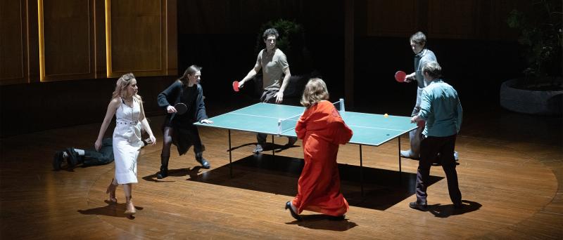 Bühnensituation. Josefine Israel, Henni Jörissen, Pascal Houdus, Daniel Hoevels, Samuel Weiss und Bettina Stucky haben alle Tischtennisschläger in den Händen und laufen um eine Tischtennisplatte herum. Im Hintergrund hohe Wände, die holzvertäfelt sind. Auch der Boden ist holzvertäfelt. Links am Rand liegt Paul Behren auf dem Boden.