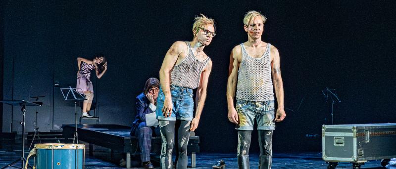 Kristof Van Boven und Nils Kahnwald stehen nebeneinander auf der Bühne. Kristofs Oberkörper neigt sich etwas in Richtung von Nils, die beiden blicken in dieselbe Richtung. Im Hintergrund sitzt Julia Wieninger auf einem Podest und beobachtet die beiden. Auf der Bühne verstreut stehen Notenständer, eine Kiste und eine Trommel. Weiter im Hintergrund tanzt Maria Carolina Vieira.