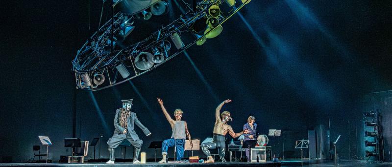 Nils Kahnwald und Kristof Van Boven stehen nebeneinander hinter einem kniehohen Podest und stellen beide den rechten Fuß darauf. Sie strecken ihre Arme nach oben und neigen sich zur Seite. Kristof Van Boven trägt eine künstliche lange gebogene Nase. Maria Carolina Vieira steht breitbeinig auf dem Podest und trägt lederne Knieschoner, eine weite schwarze Uniform mit Mütze und eine weiße Maske. Im Hintergrund stehen verteilt Notenständer und Julia Wieninger neben einer Trommel. Über der Szene hängt schräg ein