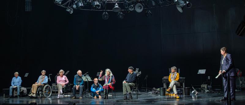 Alle sieben Zeitzeug*innen der Inszenierung sitzen nebeneinander auf der Bühne. Eine Person mittig hat eine Hand gehoben und Mittel- und Zeigefinger gespreizt. Alle schauen zu ihr. Rechts steht Julia Wieninger an einem Mikrofon, Kristof Van Boven sitzt zu den Füßen der sprechenden Person und schaut sie an. Über der Szene hängt eine große runde Stahlkonstruktion aus vielen unterschiedlich großen alten trichterförmigen Lautsprechern, Scheinwerfern und Ventilatoren.