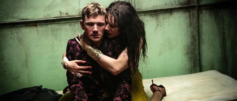 Paul Behren sitzt auf einer schmutzigen gelblichen Matratze vor einer schmutzig-rostigen Wand. Lilith Stangenberg kniet hinter ihm mit gelber Hose, goldenem Armreif und schwarzen Absatz-Schuhen. Sie hat ihre Arme um Paul Behren gelegt und drückt ihr Gesicht an seine linke Wange.