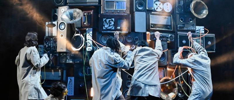 Aus mehreren Stereo- und Musikanlagen, Musikboxen und Grammophonen ist ein großer Gerätehaufen aufgebaut, vor dem fünf Personen stehen, die an den Anlagen rumdrehen.