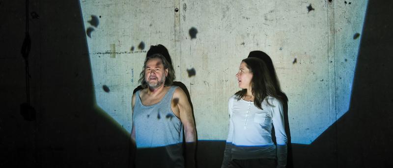 Bühnensituation. Markus John und Ute Hannig stehen vor einer Betonwand. In einem eckigen Lichtstrahl werden sie beleuchtet. Außerhalb davon ist es dunkel. Markus trägt ein hellgraues Unterhemd und schaut in die Kamera. Ute trägt ein weißes langärmliges Shirt und schaut Markus an.