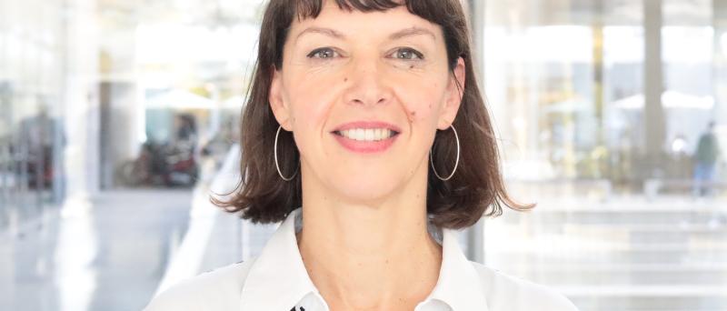Prof. Dr. Elke Schwarz steht in einem Innenraum vor einer großen Fensterfront. Sie trägt schulterlange dunkle Haare, silberne Ohrringe und eine schwarz-weiß gestreifte Bluse mit roten Details.