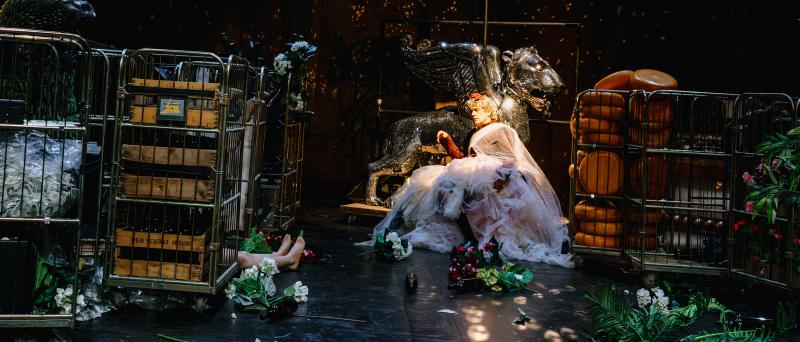 Bühnensituation. Christoph Jöde sitzt mit hellblonden Haaren und einem großen bauschigen Tüllkleid auf einem Stuhl in der Mitte der Bühne. Er lehnt an einem Requisitenwagen, in dem eine große Tierfigur aus einem Karussell steht, die das Licht in alle Richtungen reflektiert, wie eine Diskokugel. Er hat die Augen geschlossen. Um ihn herum stehen weitere Requisitenwagen mit ganz verschiedenen Requisiten darin. Auf dem Boden sind künstliche Schnittblumen verteilt.