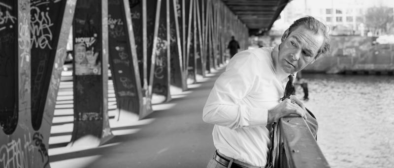 Außenansicht. Schwarz-Weiß-Bild. Mirco Kreibich steht leicht über ein Geländer gebeugt auf einer Brücke über einem Fluss. Links von ihm sind die Brückenaufbauten im Bild. Mirco dreht sich leicht über seine rechte Schulter und schaut hinter sich. Er trägt ein weißes Hemd, eine Krawatte, eine helle Hose mit Gürtel und einen großen Ring am rechten Zeigefinger. Seine Jacke hat er über das Geländer gelegt. Rechts im Bild ist das Wasser des Flusses im Bild.