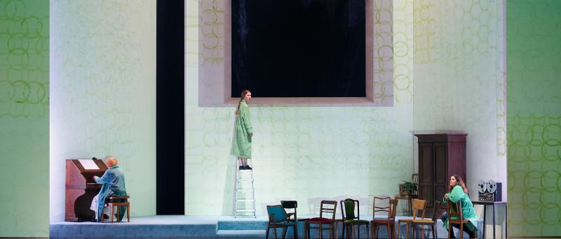 Bühnensituation: Das Bühnenbild besteht aus hohen weißen Wänden auf denen grüne überlappende Kreise aufgemalt sind. Die mittlere Wand hat eine große rechteckige Öffnung, dahinter ist es schwarz. Ilse Ritter sitzt mit dem Rücken zum Publikum links an einem Klavier. Mittig steht Alberta von Polnitz auf einer Leiter. Rechts sitzt Julia Wieninger auf einem Stuhl. Alle drei tragen grün-blaue Bademäntel. Mehrere Stühle sind auf der Bühne verteilt. Rechts ein Holzschrank sowie ein Tonbandgerät auf einem Tisch.