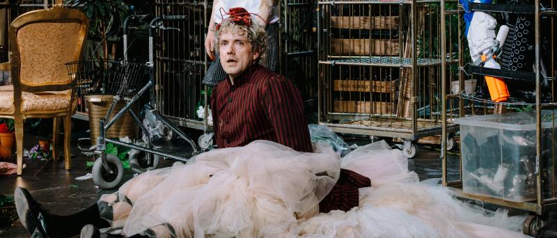 Bühnensituation. Christoph Jöde sitzt auf dem Boden und trägt einen großen Tüllrock und eine rot-schwarz gestreifte Jacke und schwarze Stiefel. Hinter ihn stehen zahlreiche Wagen mit Requisiten drauf. Links hinter ihm steht ein goldener Stuhl. Direkt hinter ihm steht eine weitere Person. Ihr Gesicht ist nicht zu sehen, man sieht nur ihr Arme, ihre Beine und einen Teil ihres Oberkörpers. Sie trägt ein weißes T-Shirt und eine gestreifte kurze Hose.