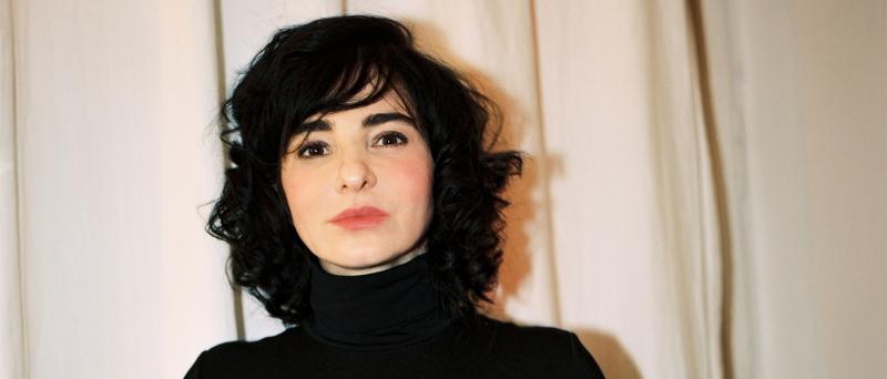 Mely Kiyak steht vor einem hellen Hintergrund in einem Innenraum. Sie trägt ein schwarzes hochgeschlossenes Oberteil, schulterlange schwarze Haare und schaut in die Kamera.