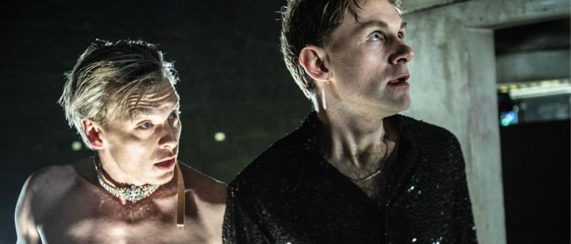 Franz Pätzold und Paul Behren stehen nebeneinander und gucken mit leicht geöffnetem Mund und schauen beide in dieselbe Richtung. Franz Pätzold ist leicht zu Paul Behren gedreht. Franz Pätzold ist Oberkörperfrei und trägt eine mit Diamanten bestückte Kette. Paul Behren trägt eine leicht durchsichtige schwarze Glitzer Bluse.