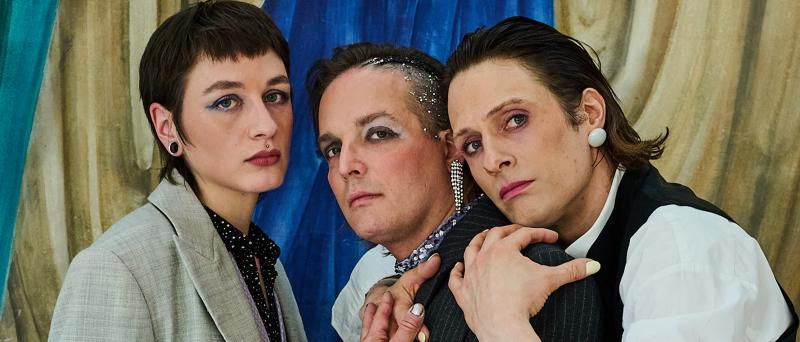 Juno Peter, Micha Krause und Less Brandes-Hell stehen eng nebeneinander und schauen in die Kamera. Less hat ihren Kopf und die Arme um Michas Schultern gelegt, Micha berührt ihre Hand. Sie stehen vor einem gemalten gelben Vorhang.