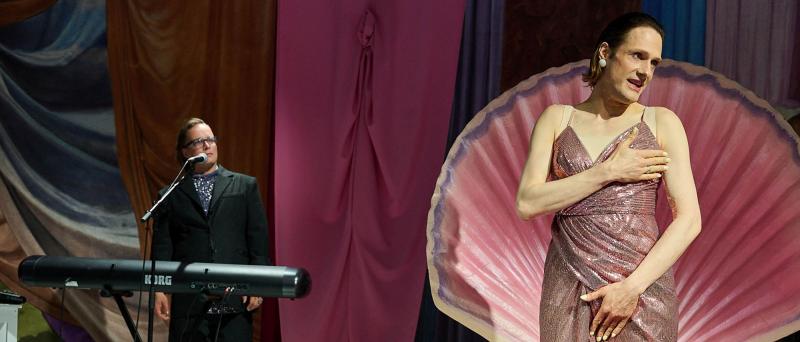 Bühnensituation: Less Brandes-Hell steht in einer rosafarbenen aufgeklappten Muschel. Sie trägt ein rosa Glitzerkleid, hält sich eine Hand vor die Brust und eine Hand vor den Schritt. Sie hat den Mund leicht geöffnet und schaut nach rechts. Im Hintergrund steht Micha Krause hinter einem Keyboard und am Mikro. Sie trägt einen schwarzen Anzug mit einem blauen Oberteil darunter sowie schwarze Stiefel. Der Hintergrund ist mit bunten Stoffen verhangen.