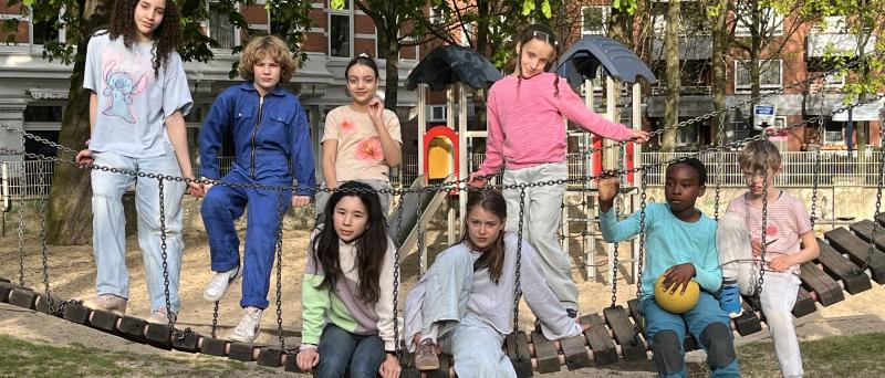 Zu sehen sind acht Kinder, die auf einer Hängebrücke eines Spielplatzes stehen und sitzen. Sie tragen unterschiedliche, bunte Kleidung und schauen in die Kamera. Im Hintergrund sind Bäume zu erkennen.