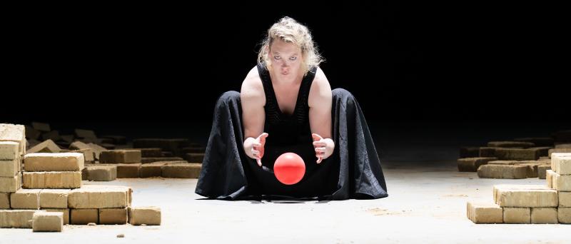 Lina Beckmann hockt in schwarzem Gewand auf einer kargen Bühne und beugt sich intensiv über eine leuchtend rote Kugel, die Hände wie beschwörend darüber ausgestreckt. Ihr Blick ist eindringlich, die Haare zerzaust. Rechts und links sind sandfarben gestapelte Ziegelsteine verteilt, der Hintergrund liegt im Dunkel. Die Szene strahlt eine bedrohliche, rituelle Atmosphäre aus.