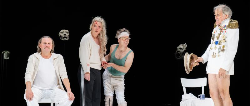 Devid Striesow, Lina Beckmann, Kristof van Boven und Sebastian Blomberg stehen auf einer weißen Bühne vor schwarzem Hintergrund mit schwebenden Masken. Links Devid Striesow in weißem Jogginganzug. Daneben Lina Beckmann und Kristof van Boven in Alltagskleidung – eine mit zerzaustem Haar und Blutspuren an der Stirn, der andere mit Stirnband und Augenklappe. Rechts steht Sebastian Blomberg in einem pompösen Militäruniform-Oberteil, roten Socken und ohne Hose, der einen Hut in der Hand hält.