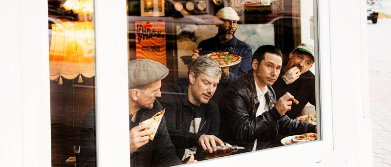 Fünf Männer sitzen in einem Restaurant hinter einer großen Fensterscheibe und essen Pizza. Einer der Männer hält eine Pizza hoch, während die anderen Pizza­stücke in der Hand halten. Die Männer tragen überwiegend dunkle Kleidung und teilweise Schiebermützen oder eine Kappe.