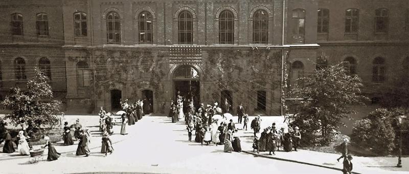 Historische Schwarzweißaufnahme des Universitätsklinikums Hamburg-Eppendorf (UKE). Vor einem repräsentativen Klinikgebäude mit großen Rundbogenfenstern stehen und gehen zahlreiche Menschen, darunter viele Frauen und Kinder. Die Aufnahme zeigt das UKE in historischer Zeit.