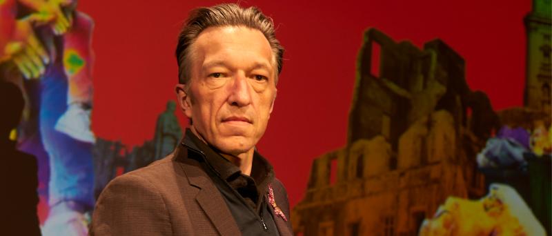 Porträt des Autors und Dramatikers Lukas Bärfuss in einem dunklen Sakko und schwarzem Hemd vor einer roten Kulisse mit einer düsteren, ruinösen Gebäudedarstellung und farbigen Figuren an den Bildrändern.