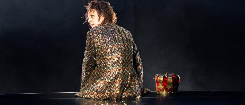 Lina Beckmann sitzt mit dem Rücken zur Kamera auf der Rückseite der dunklen Bühne, in einem funkelnden Blazer aus Gold, Rot und Silber. Den Kopf dreht sie halb nach hinten. Neben ihr liegt eine rot-goldene Krone auf dem Boden.