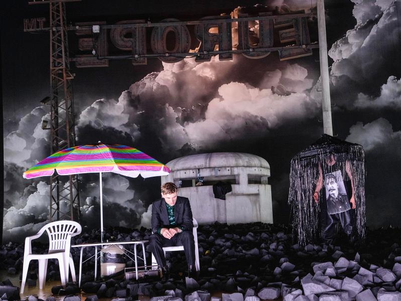 Der Schauspieler Paul Behren sitzt auf einem weißen Plastikstuhl unter einem bunten Sonnenschirm. Um ihn überall schwarze Kohle um ihn herum. Im Hintergrund dunkle Wolken am Himmel und ein gigantisches Schild, das verkehrt herum steht und von der anderen Seite den Schriftzug "Europe" zeigt.