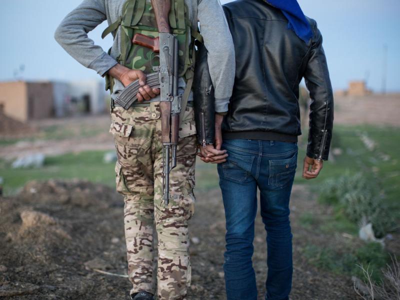 Ankündigungsmotiv "Über Grenzen gehen": Zwei Männer von hinten, sie halten sich an den Händen, einer trägt Tanrkleidung und eine Waffe auf dem Rücken