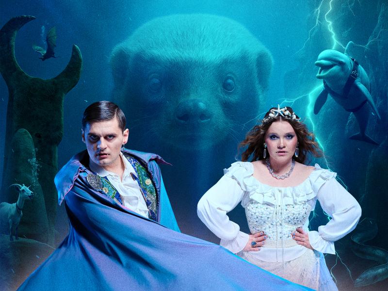 Ein Mann in einem blauen Umhang und Vampirzähnen und eine Frau in einem altmodischen weißen Kleid schauen in die Kamera. Im Hintergrund eine Unterwasserwelt, ein Delfin und der Kopf eines großen Otters.