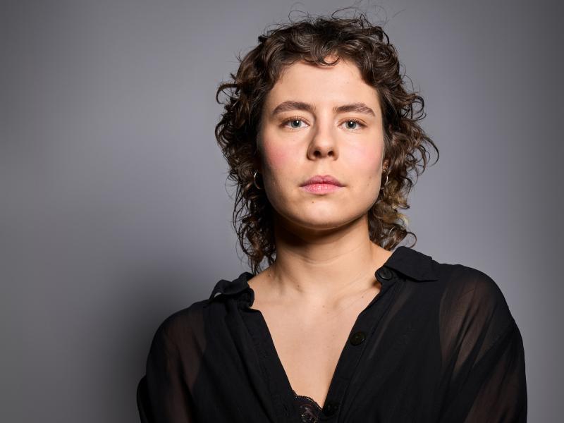 Die Schauspielerin Anastasia Lara Heller steht vor einem grauen Hintergrund. Sie trägt dunkelbraue Locken, ein schwarzes Oberteil und schaut in die Kamera.