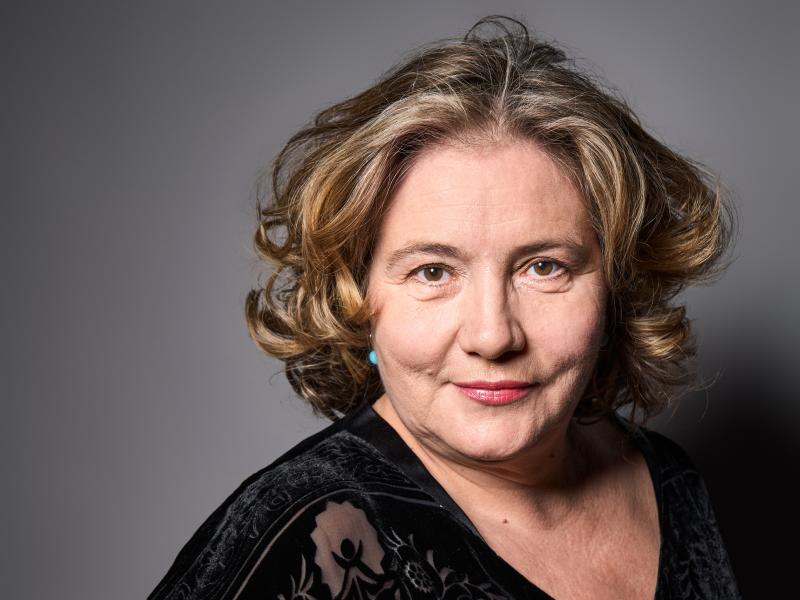 Die Schauspielerin Bettina Stucky steht vor einem grauen Hintergrund. Sie trägt dunkelblonde Locken, eine schwarze Bluse und schaut in die Kamera