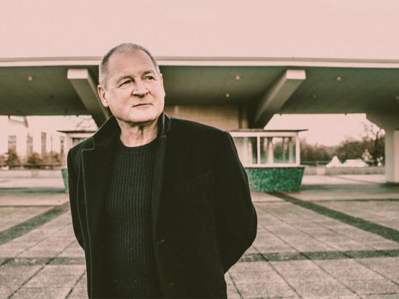 Der Schauspieler Burghart Klaußner steht im Freien. Er schaut leicht an der Kamera vorbei und trägt eine schwarze Jacke. Im Hintergrund ein Gebäude, bei dem es sich um eine alte Tankstelle handeln könnte.