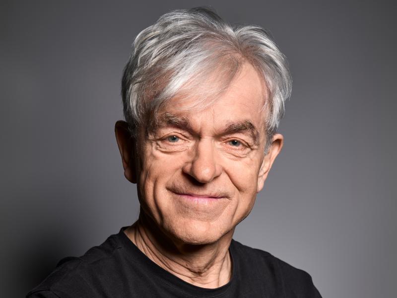 Der Schauspieler Michael Weber steht vor einem grauen Hintergrund. Es trägt hellgraue kurze Haare, ein schwarzes T-Shirt und lächelt in die Kamera.
