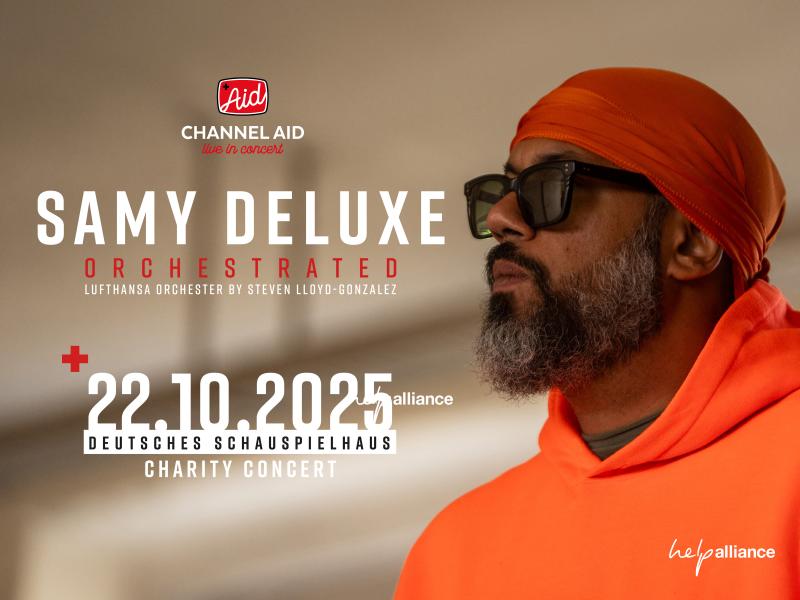 Der Künstler Samy Deluxe in orangem Pullover, roter Mütze und dunkler Sonnenbrille vor einem hellen, leicht unscharfen Hintergrund.