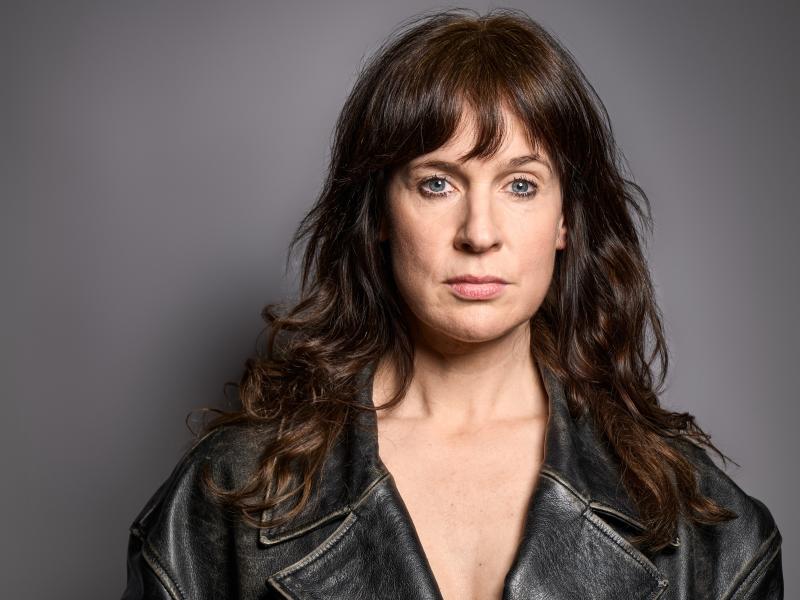 Die Schauspielerin Sandra Gerling steht vor einem grauen Hintergrund. Sie trägt ihre dunkelbraunen Haare offen. Sie trägt eine schwarze Lederjacke und schaut in die Kamera.