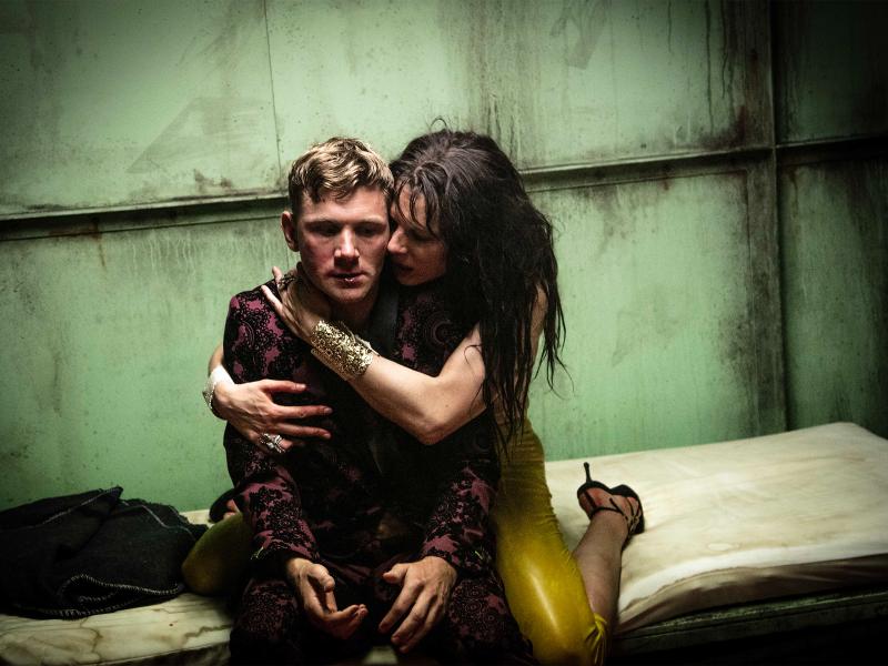 Paul Behren sitzt auf einer schmutzigen gelblichen Matratze vor einer schmutzig-rostigen Wand. Lilith Stangenberg kniet hinter ihm mit gelber Hose, goldenem Armreif und schwarzen Absatz-Schuhen. Sie hat ihre Arme um Paul Behren gelegt und drückt ihr Gesicht an seine linke Wange.